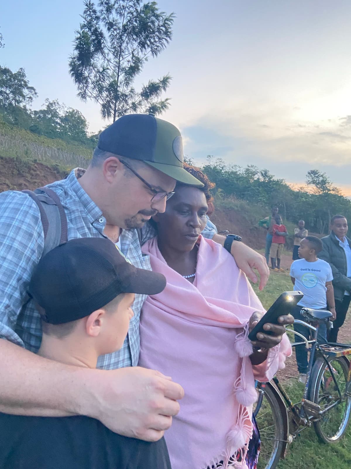 A farmer and two visitors looking together at a mobile phone screen at the edge of a field in the evening light.