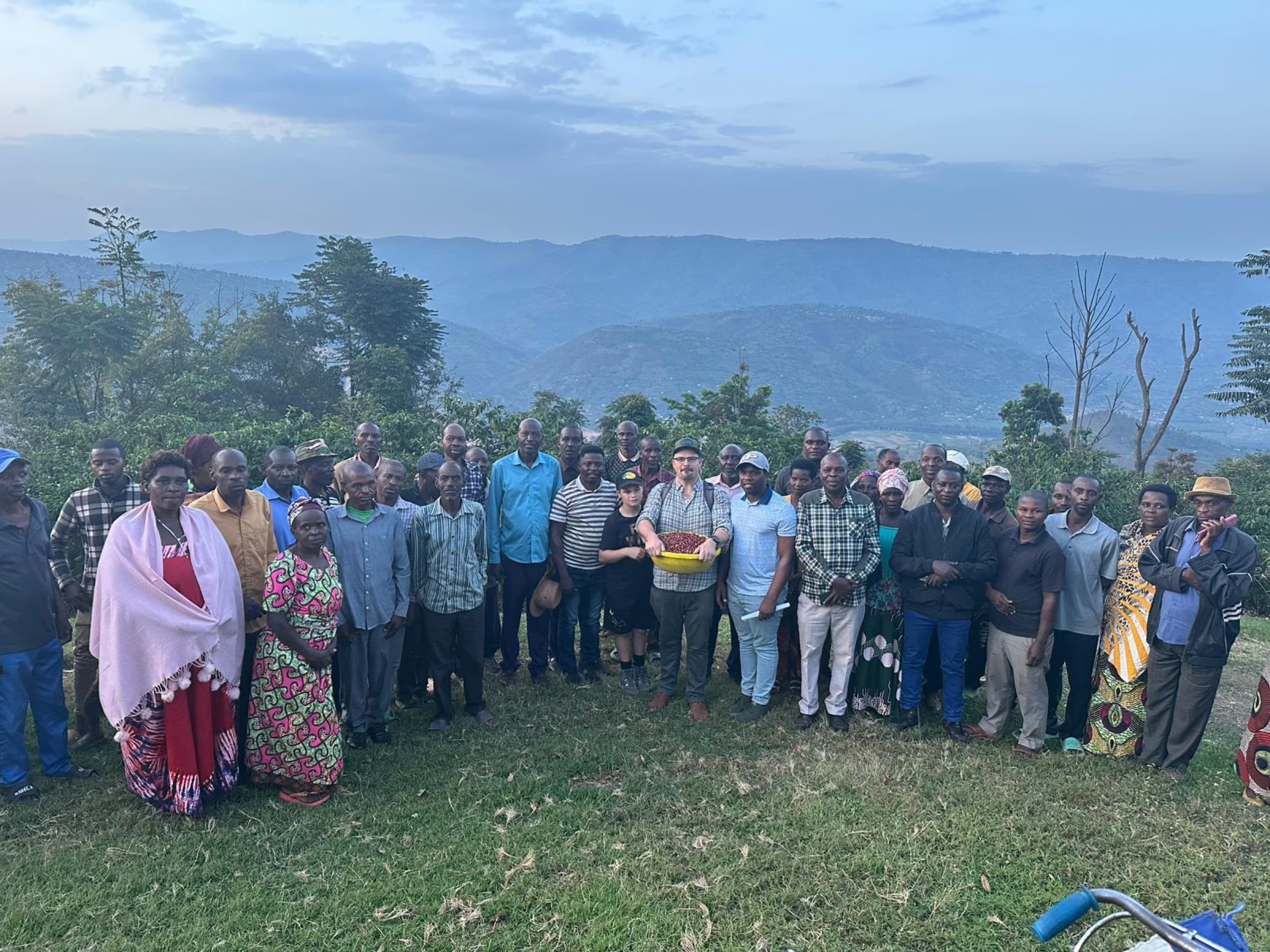 A large group of farmers and Enable Rwanda staff standing together on a hillside at dusk, with terraced fields and mountains behind them.