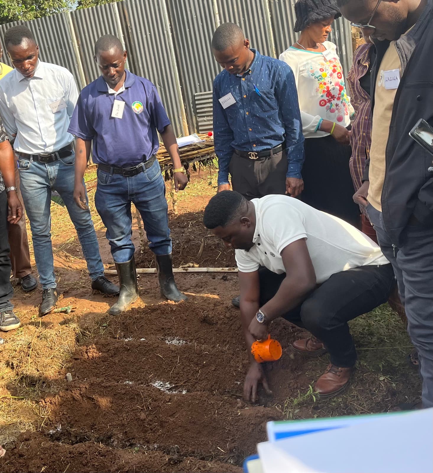 A trainer kneeling in a demonstration plot, pouring fertiliser into a planting hole while a circle of farmers looks on.