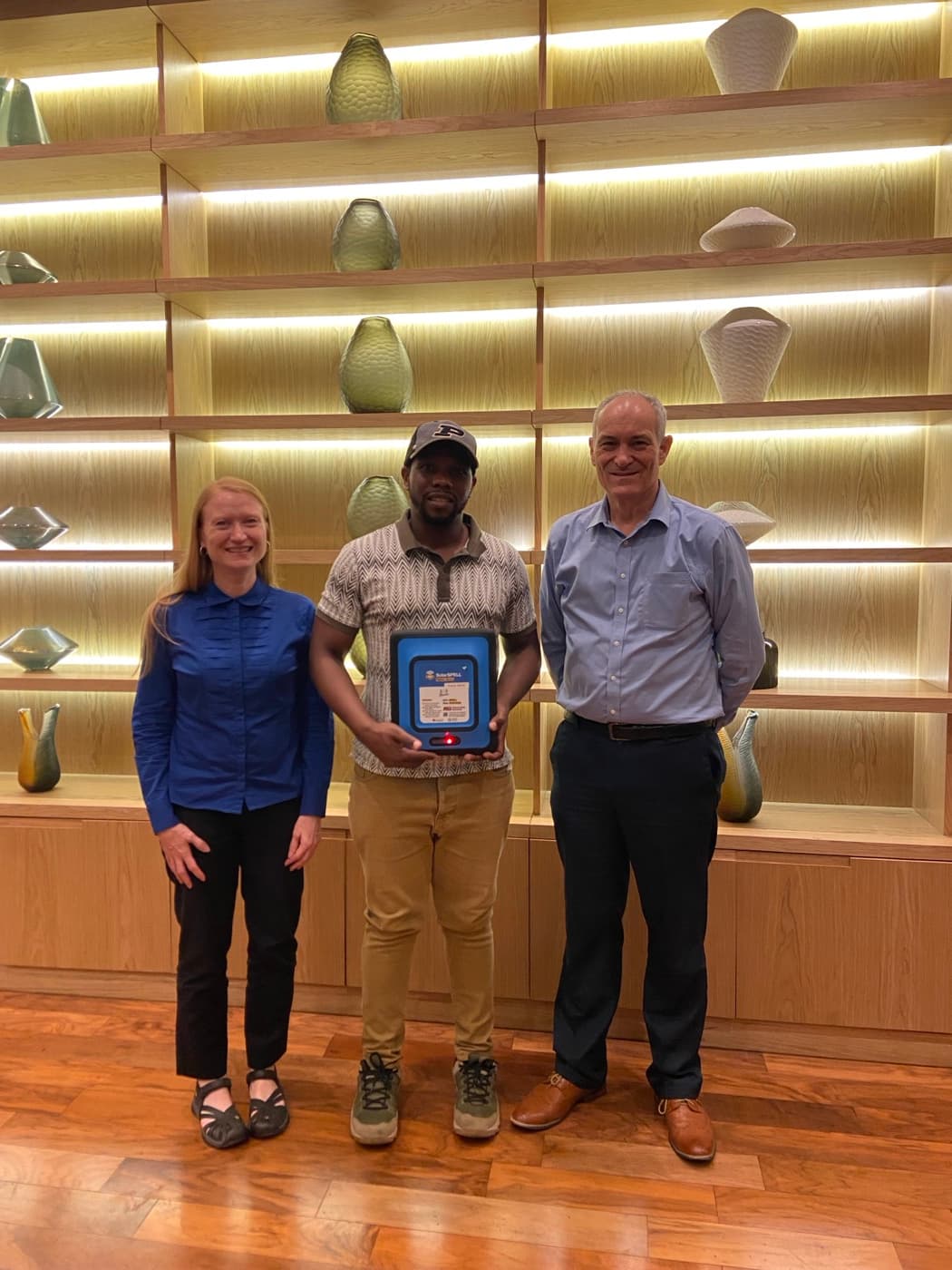 Three people standing together indoors, one holding a framed plaque marking a new partnership.