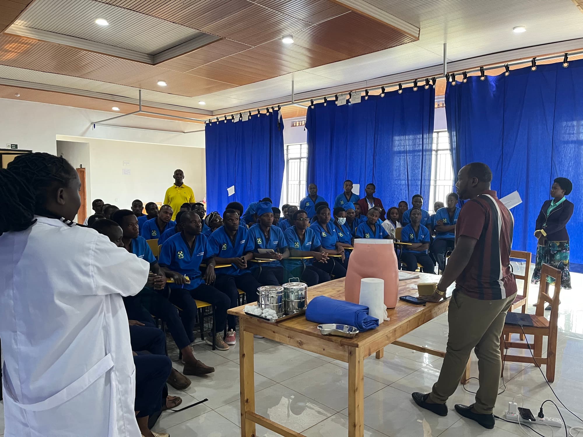 A facilitator leading a practical session for a room of students in blue overalls seated around a demonstration table.