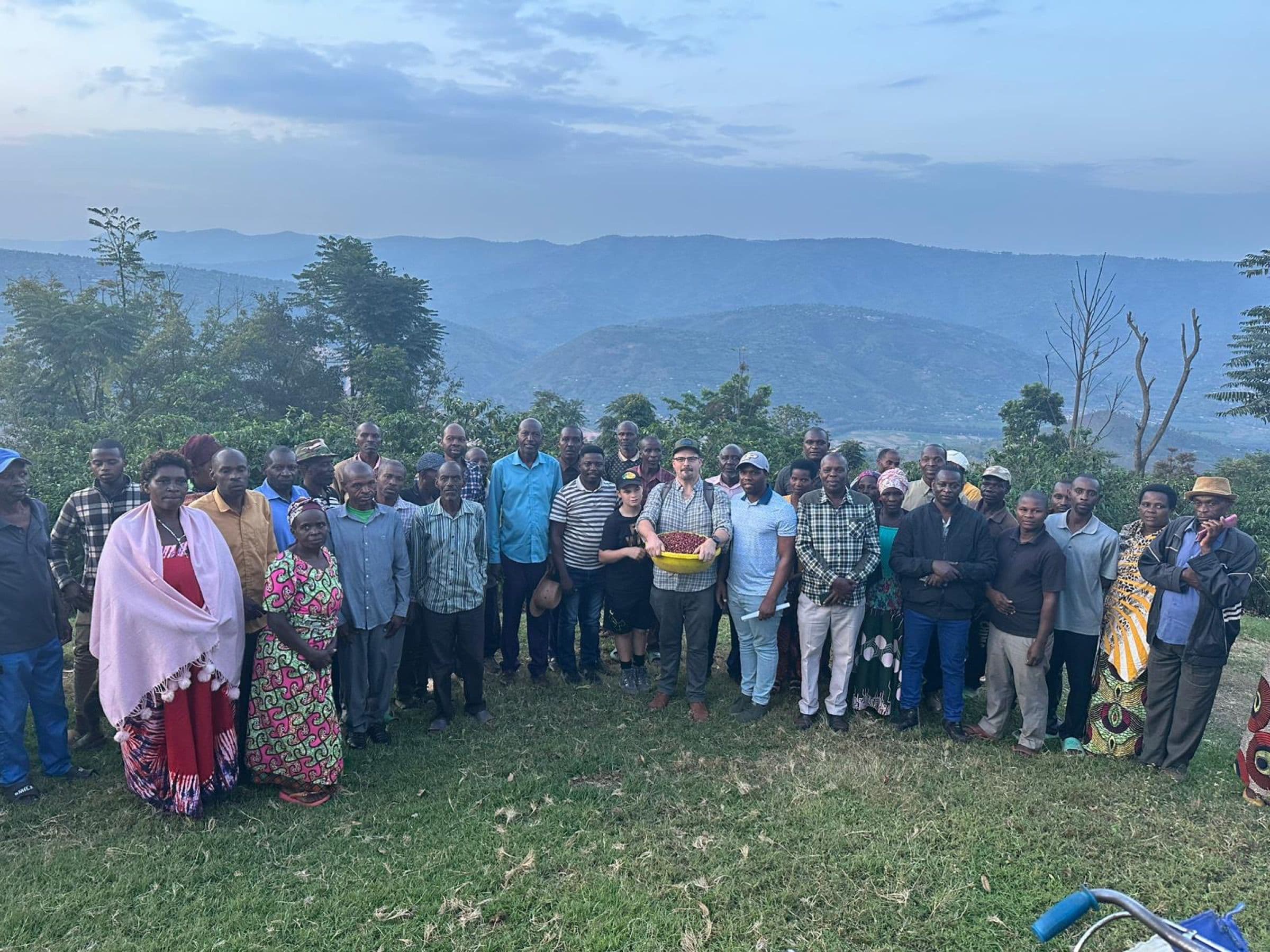 A large group of farmers and ENABLE Rwanda staff standing together on a hillside at dusk, with terraced fields and mountains behind them.